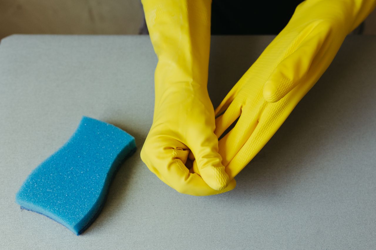 Close-up view of hands in yellow gloves beside a blue sponge on a gray surface.
