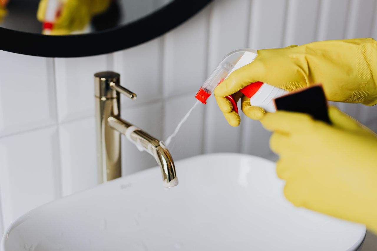 services-07 From above anonymous housekeeper in yellow rubber gloves spraying cleaner to chrome water faucet in bathroom