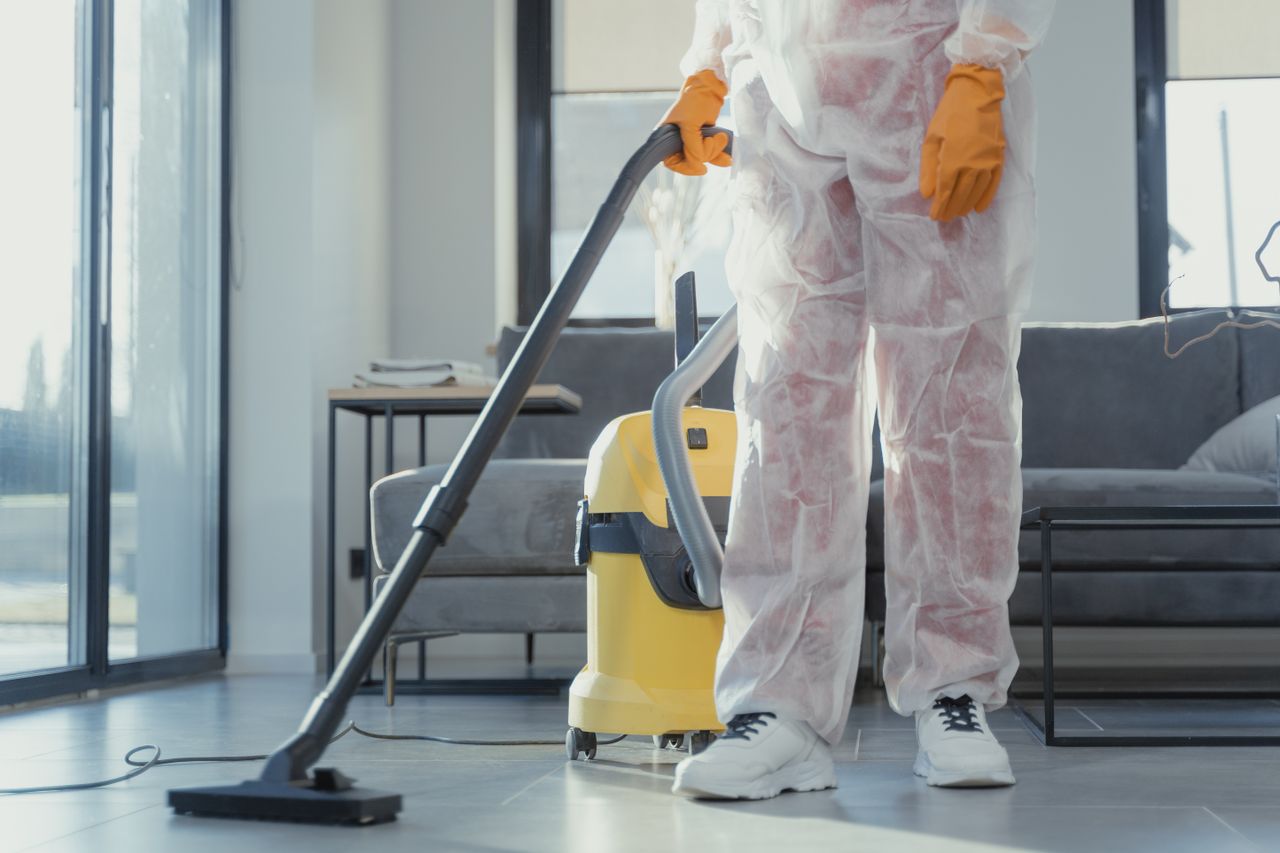 A cleaner wearing protective coveralls and gloves vacuuming a contemporary living room.